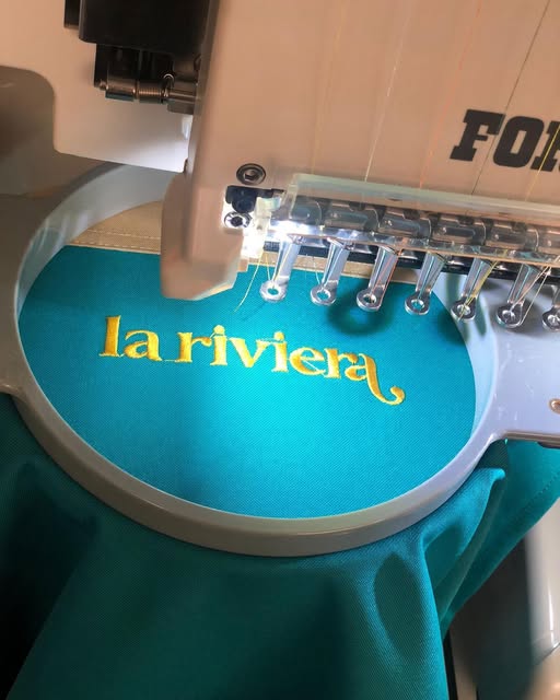 Logo bordado en hilo dorado sobre tela turquesa en la máquina
